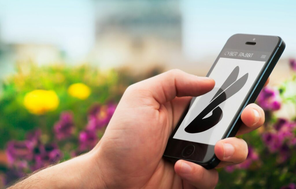 Close-up of a hand holding a smartphone with a blurred floral background, showing a logo on screen.